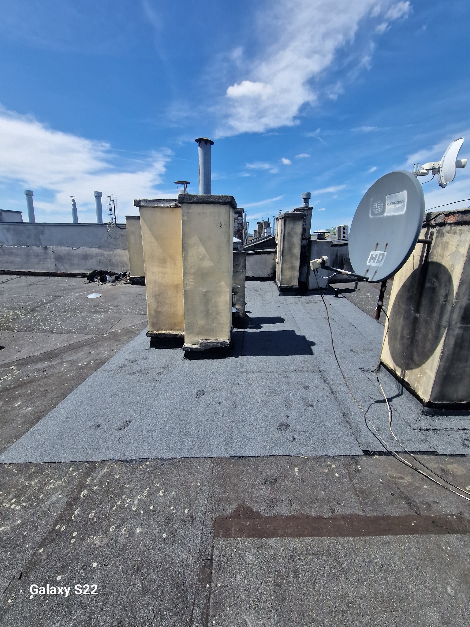 Fragment dachu płaskiego po naprawie, widoczne nowe pokrycie bitumiczne, w tle kominy wentylacyjne i antena satelitarna.