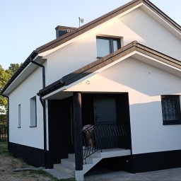 ŁASZ - BUD Łukasz Wierzbicki - Biały dom z czarnymi akcentami, widok z zewnątrz, z widocznym fragmentem osoby na ganku i samochodem w tle.