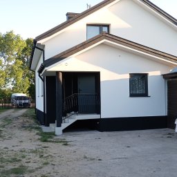 ŁASZ - BUD Łukasz Wierzbicki - Biało-czarny dom jednorodzinny z podestem wejściowym, widok z boku na elewację w słoneczny dzień, z widocznym fragmentem ogrodu i zaparkowanym busem w tle.