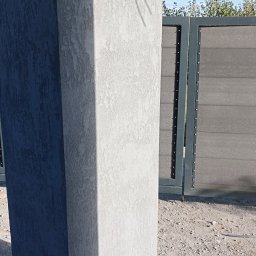 ŁASZ - BUD Łukasz Wierzbicki - Szary słup z fakturą elewacyjną, częściowo zacieniony, z widoczną bramą w tle i zwisającym kablem rzucającym cień na słup.