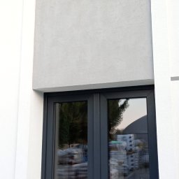 ŁASZ - BUD Łukasz Wierzbicki - Fragment elewacji budynku z oknem i drzwiami balkonowymi, wykończony jasnoszarym tynkiem strukturalnym.