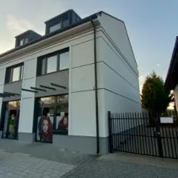 Dwupiętrowy budynek po renowacji z nową, białą elewacją, szarymi akcentami i czarnymi elementami wykończeniowymi, widoczny z zewnątrz, z salonem fryzjerskim na parterze. Po prawej stronie widoczny...