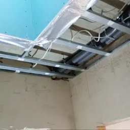 Stelaż sufitowy z profili metalowych, izolacja folią aluminiową, rury wentylacyjne w peszlu, niebieska płyta na suficie, surowe ściany z notatkami ołówkiem.