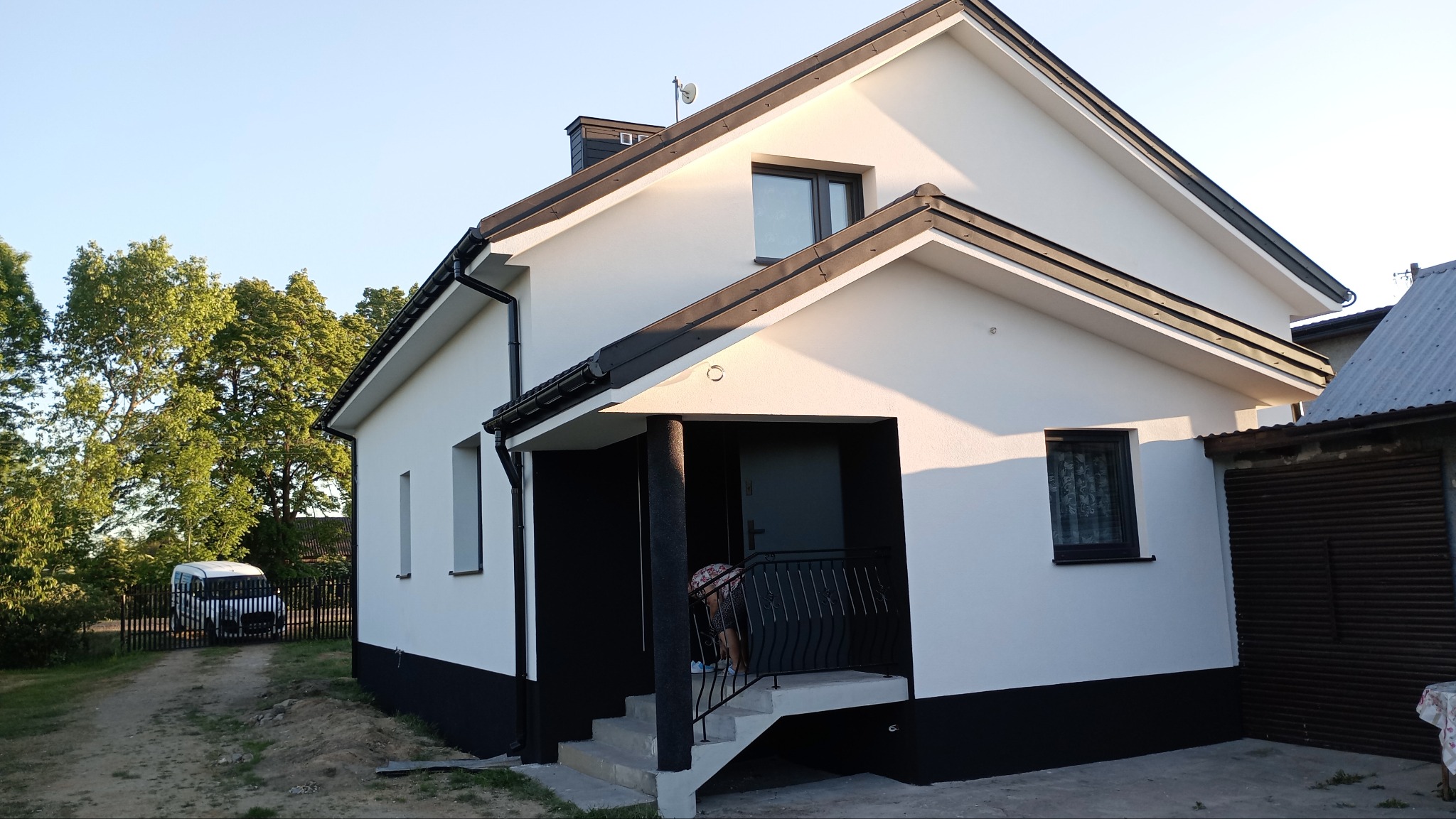 Biały dom z czarnymi akcentami, widok z zewnątrz, z widocznym fragmentem osoby na ganku i samochodem w tle.