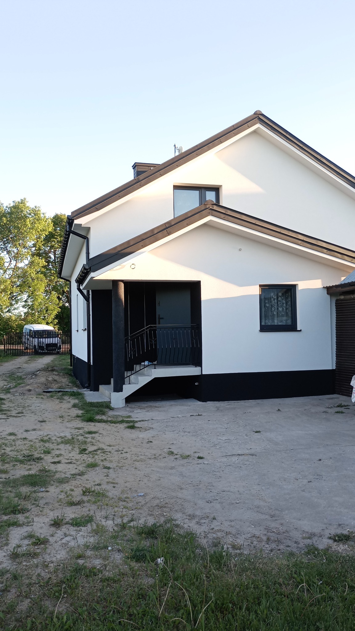 Biało-czarny dom jednorodzinny z podestem wejściowym, widok z boku na elewację w słoneczny dzień, z widocznym fragmentem ogrodu i zaparkowanym busem w tle.