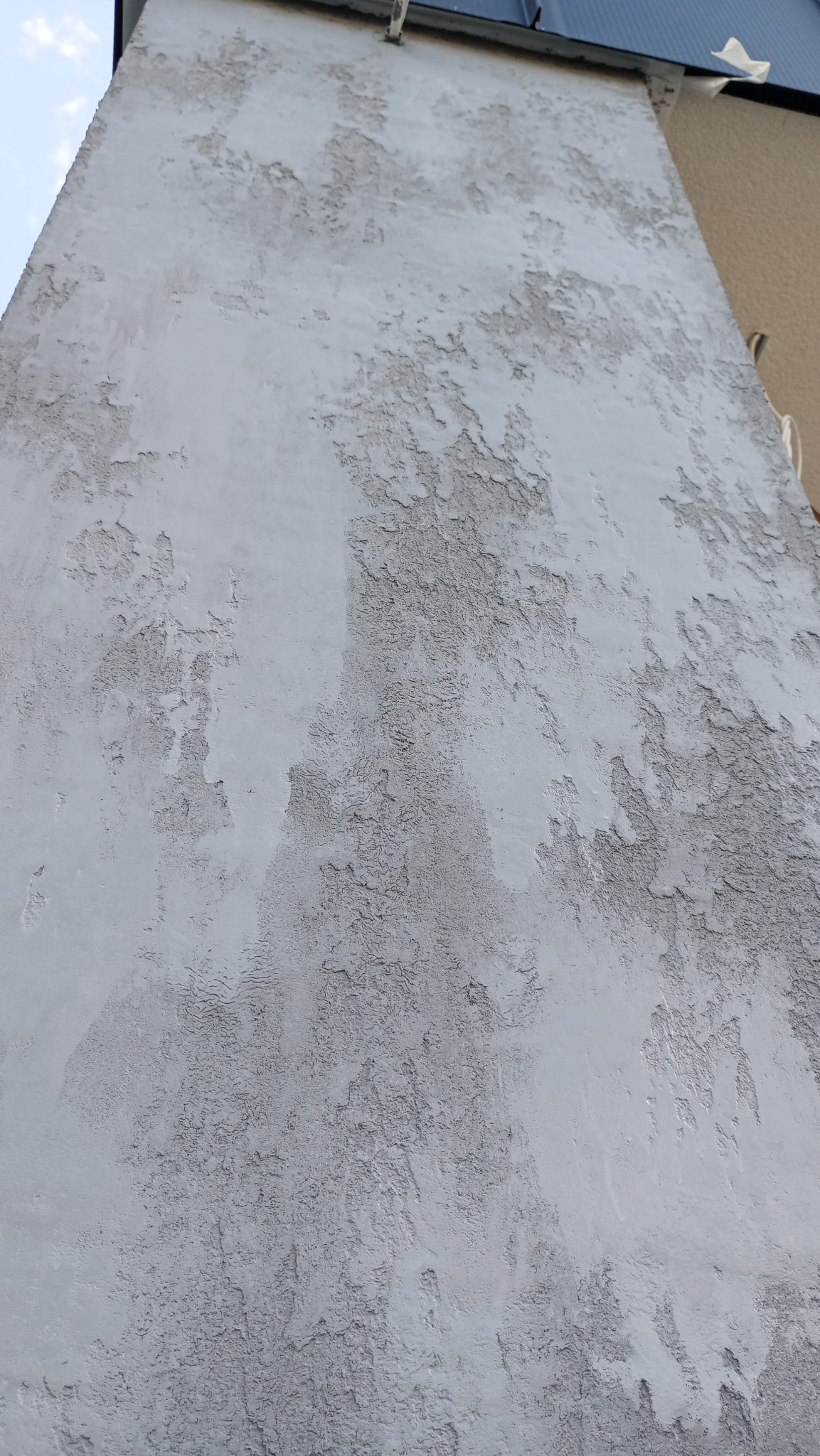 Niedokończona, szara elewacja budynku z wyraźnymi, nieregularnymi śladami po nałożonym tynku, widoczne fragmenty dachu i sąsiedniego budynku.