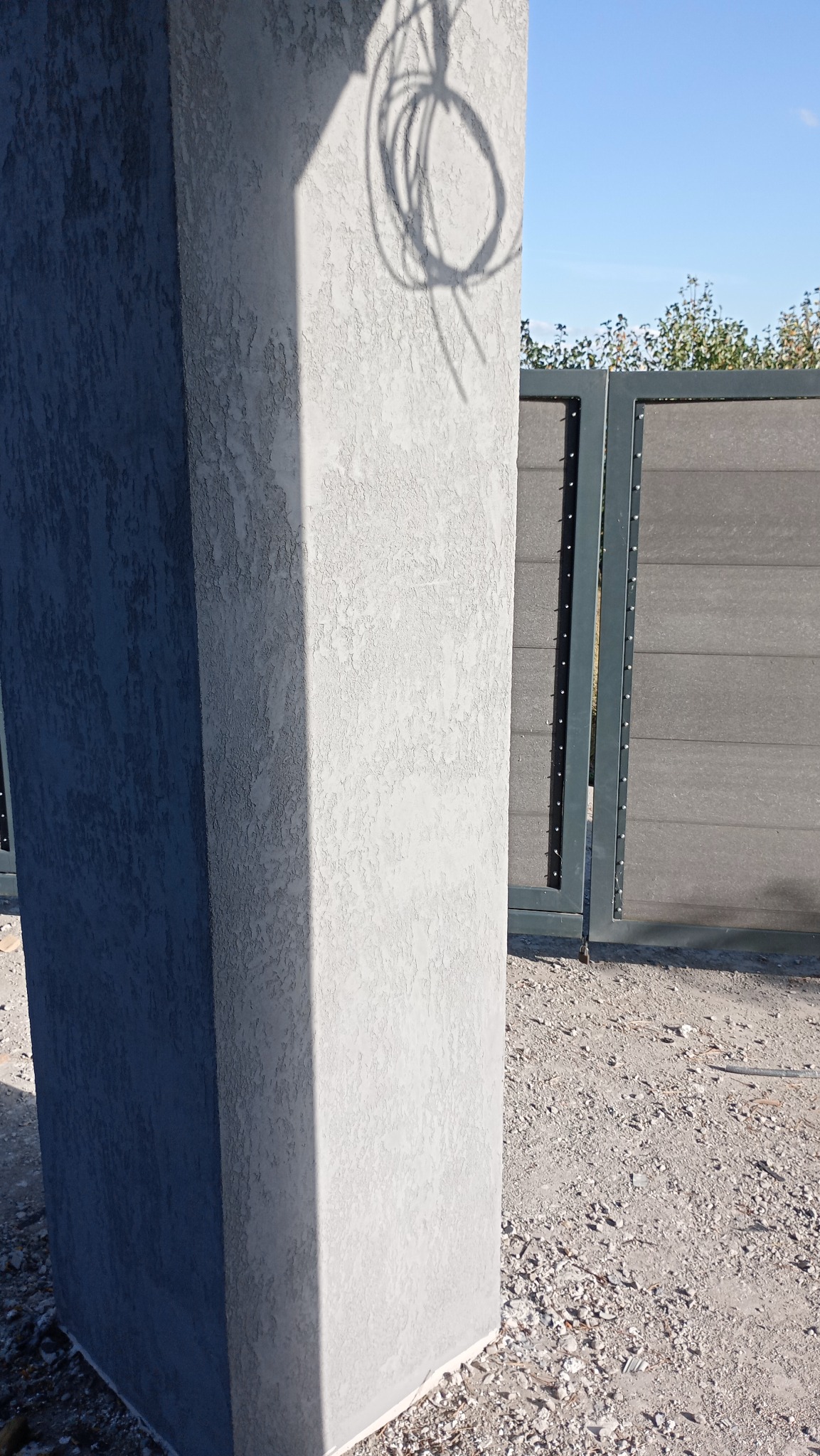 Szary słup z fakturą elewacyjną, częściowo zacieniony, z widoczną bramą w tle i zwisającym kablem rzucającym cień na słup.