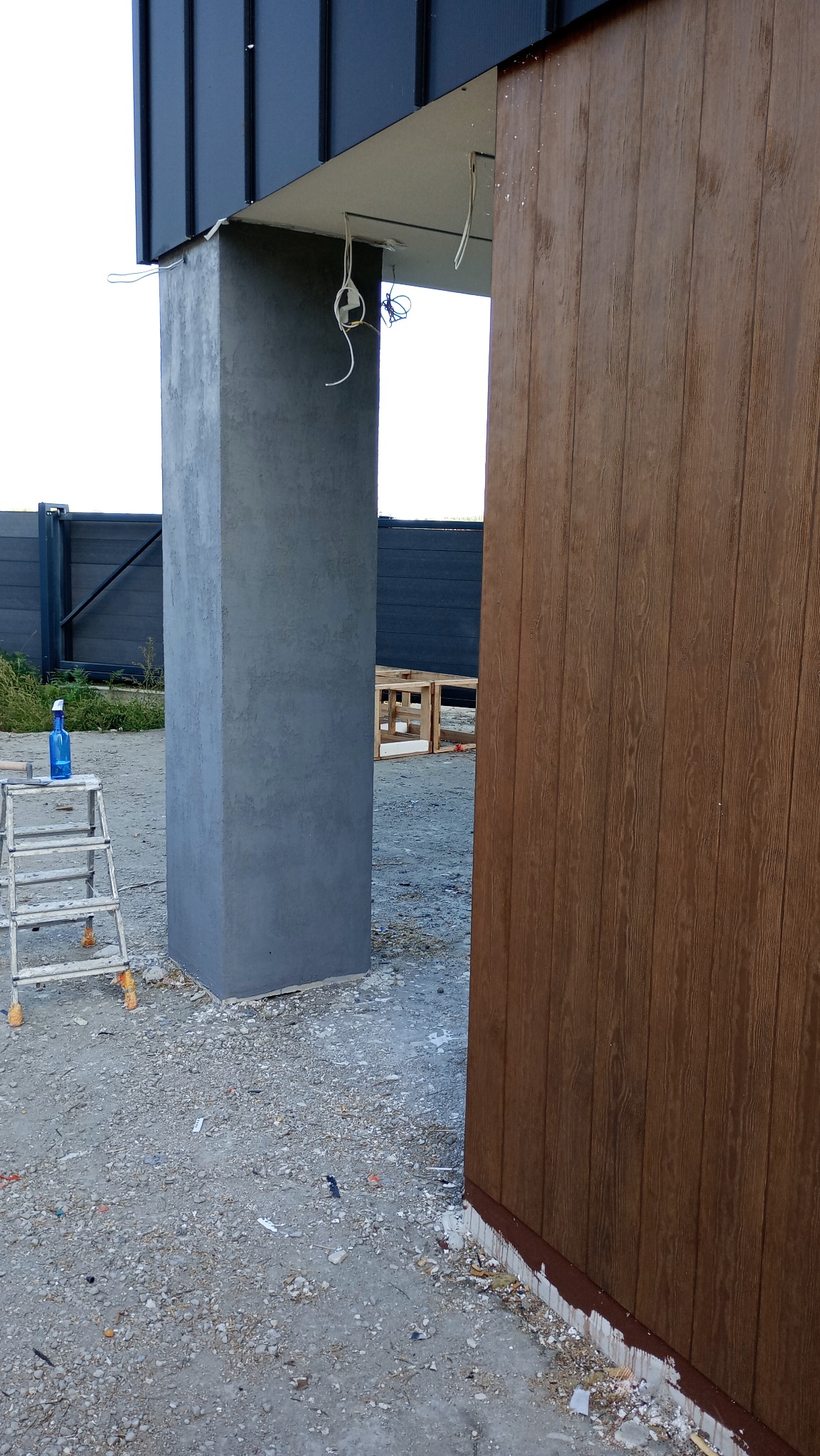 Wykończony filar z szarą strukturą obok brązowej elewacji panelowej, drabina z niebieskim płynem czyszczącym w tle, widoczne przewody elektryczne