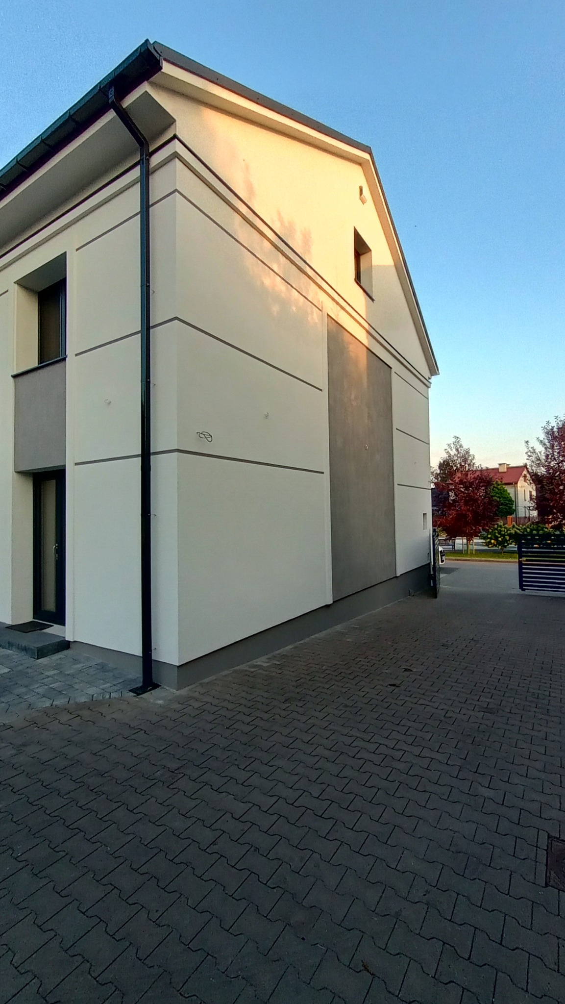 Dwukondygnacyjny dom z jasną elewacją podzieloną poziomymi szarymi pasami, czarna rynna spustowa, kostka brukowa na podjeździe, widok z boku w słoneczny dzień.