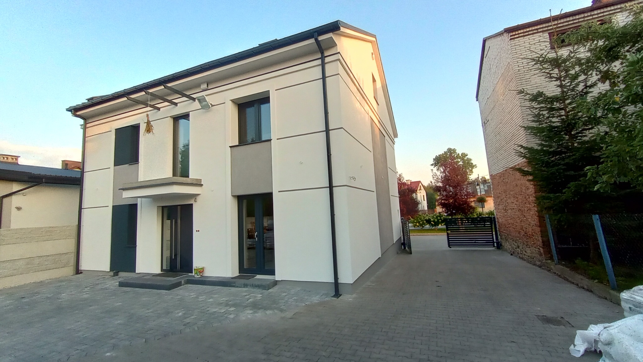 Dwukondygnacyjny budynek z nową, jasną elewacją i szarymi akcentami, widziany z poziomu brukowanego podjazdu w słoneczny dzień.