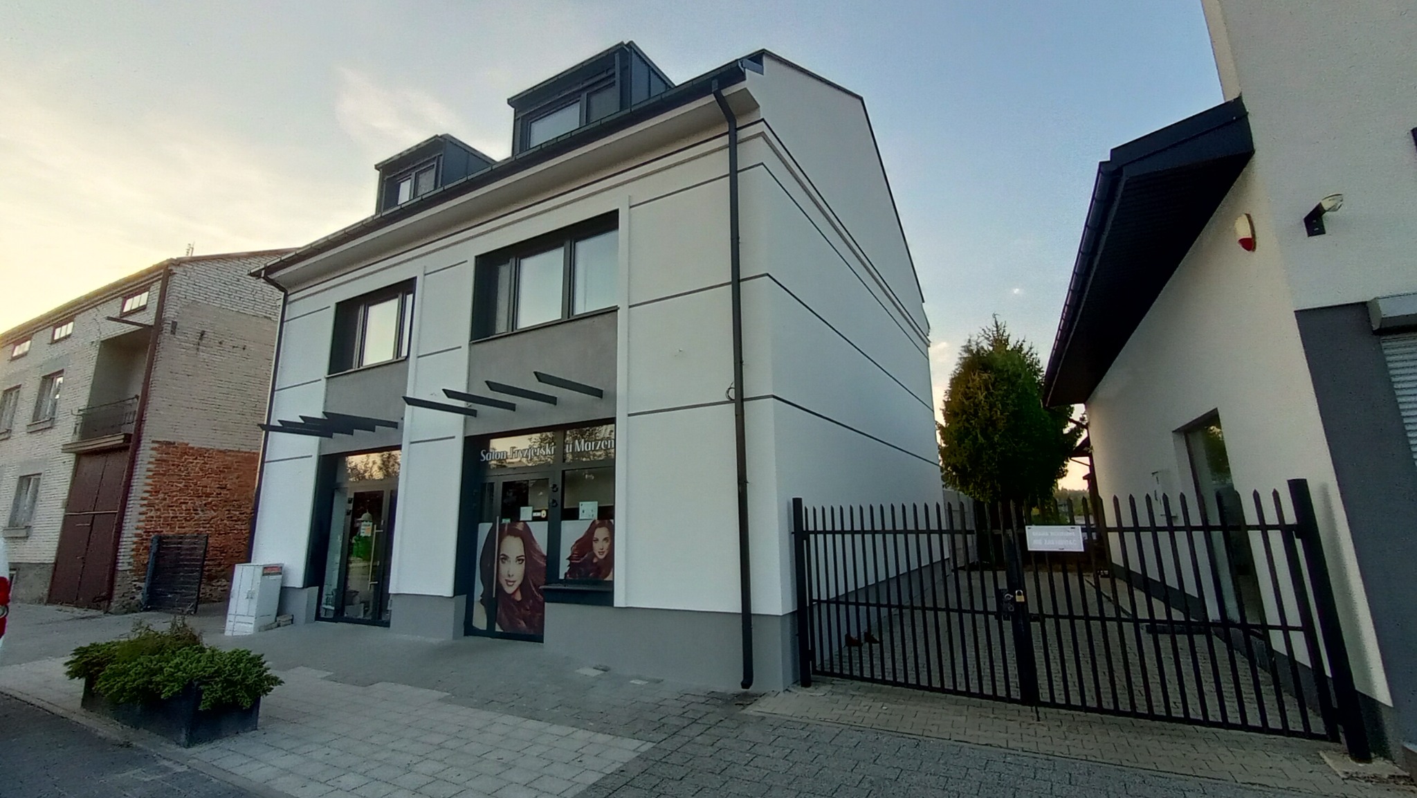 Dwupiętrowy budynek po renowacji z nową, białą elewacją, szarymi akcentami i czarnymi elementami wykończeniowymi, widoczny z zewnątrz, z salonem fryzjerskim na parterze. Po prawej stronie widoczny...