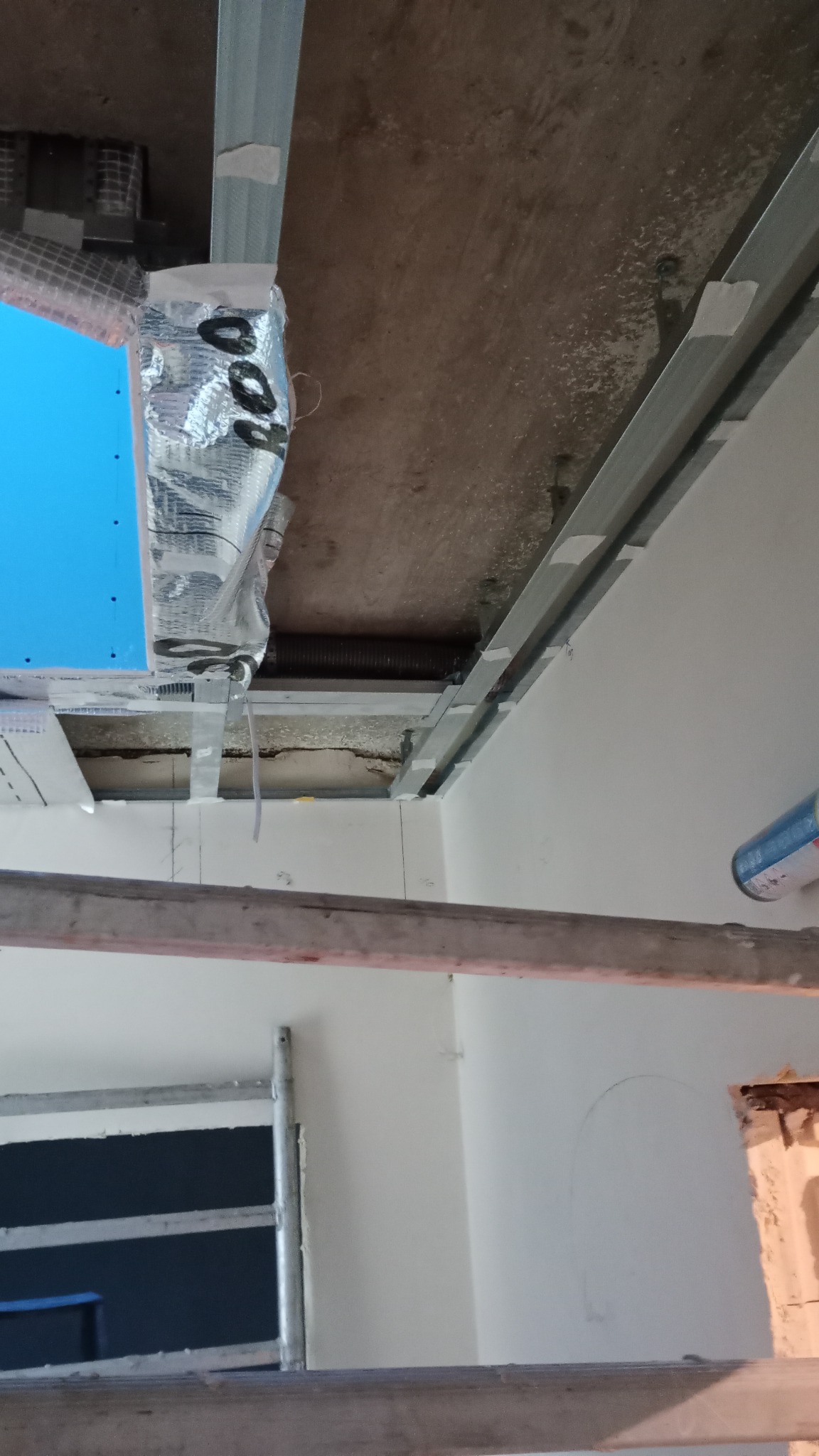 Stelaż metalowy podwieszanego sufitu z płyt, widoczne elementy instalacji i izolacji, fragment ściany z zaznaczonym łukiem, widok z rusztowania.