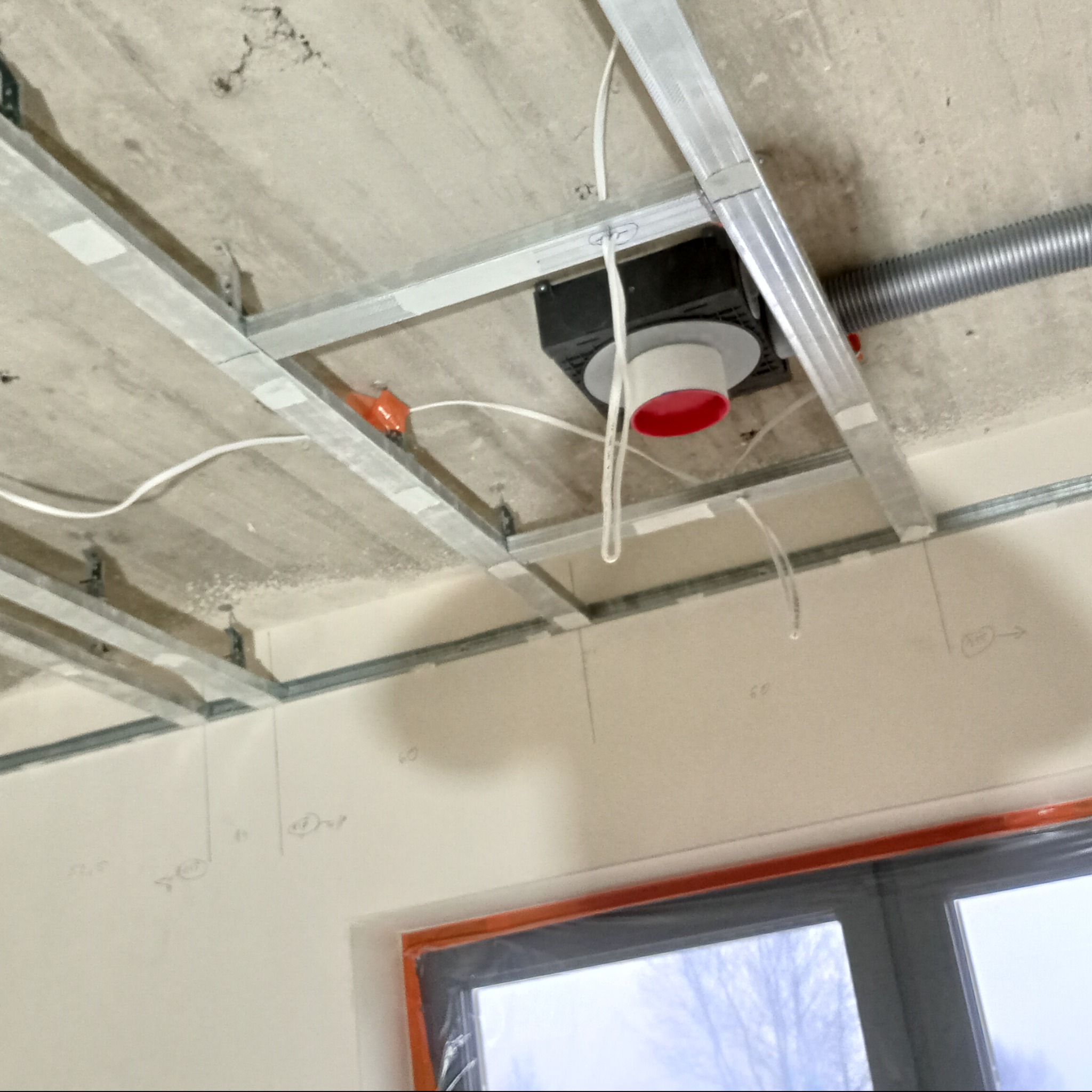 Metalowa konstrukcja sufitowa z zamontowaną wentylacją, widoczne przewody elektryczne i zaznaczenia na ścianie, okno w tle.