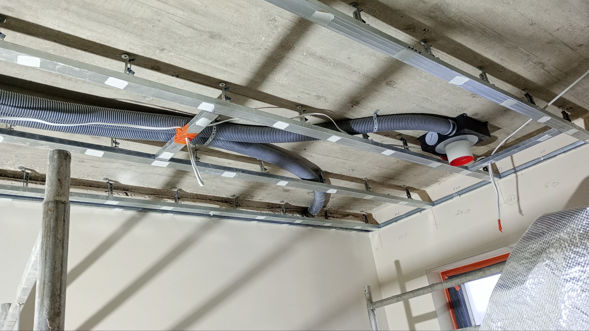 Montaż instalacji wentylacyjnej pod sufitem podwieszanym, widoczne rury wentylacyjne, profile metalowe, mocowania i fragment ściany z rusztowaniem.