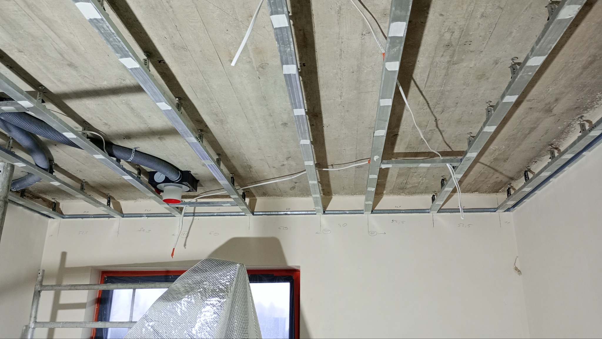 Montaż metalowej konstrukcji sufitu podwieszanego z widocznymi elementami wentylacji i przewodami elektrycznymi, częściowo zakryty folią ochronną fragment okna w tle.