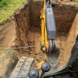 Karol Kowalski - Widok z kabiny żółtej koparki JCB podczas wykopywania głębokiego, kwadratowego dołu w ziemi na trawiastym terenie z widocznym ogrodzeniem i domem w tle.