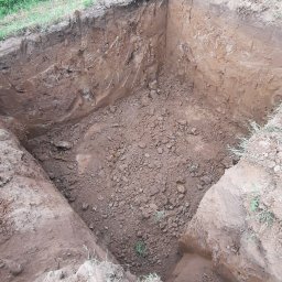 Karol Kowalski - Wykopany dół w ziemi z widocznymi ścianami z gliny i ziemi, częściowo zasypany gruzem, obok rosną trawy, widoczne buty pracownika.