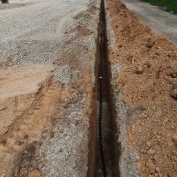 Karol Kowalski - Wykop w ziemi z ułożonym kablem, widok z góry, w tle plac budowy i pochmurne niebo.