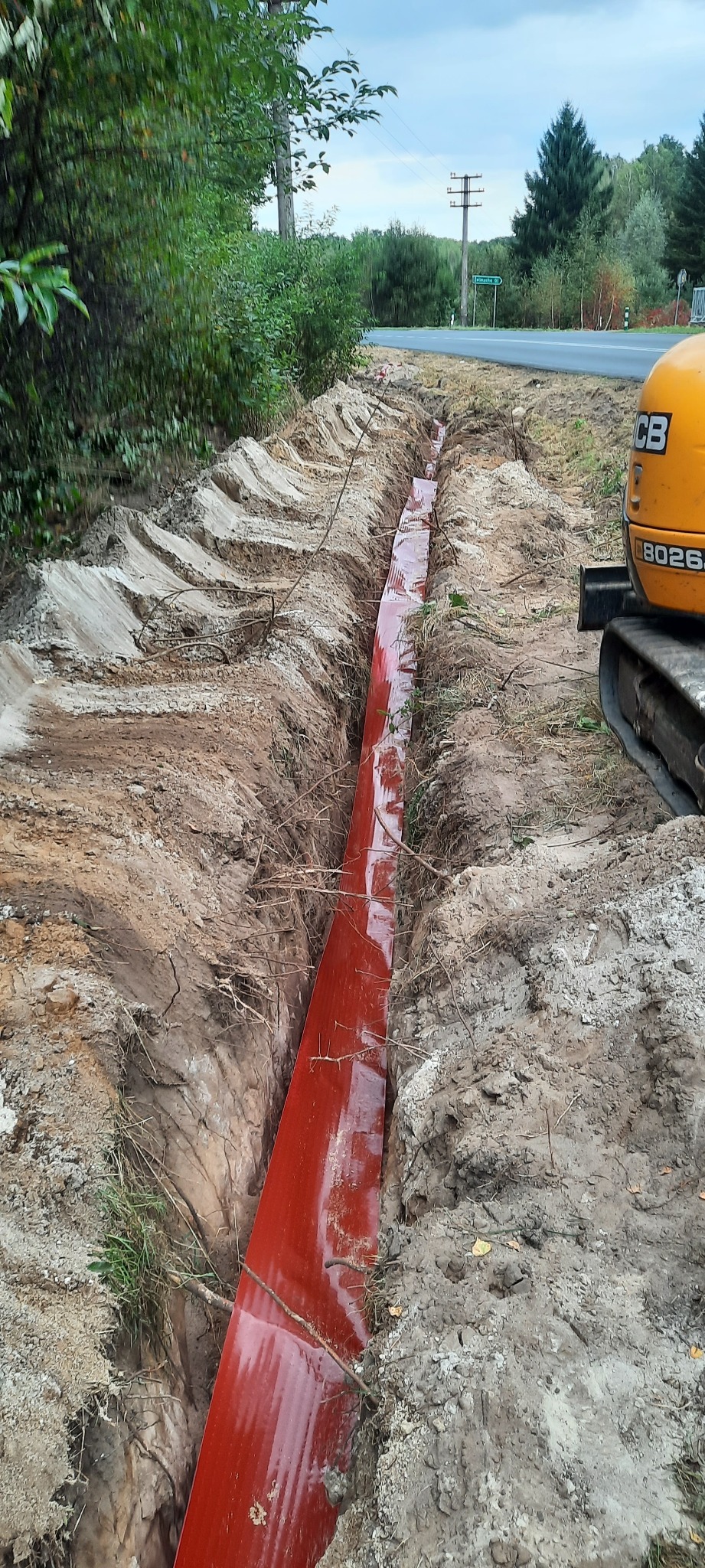 Wykopany rów w ziemi z ułożoną czerwoną rurą kanalizacyjną, widoczny fragment koparki na tle krajobrazu z drogą i drzewami.