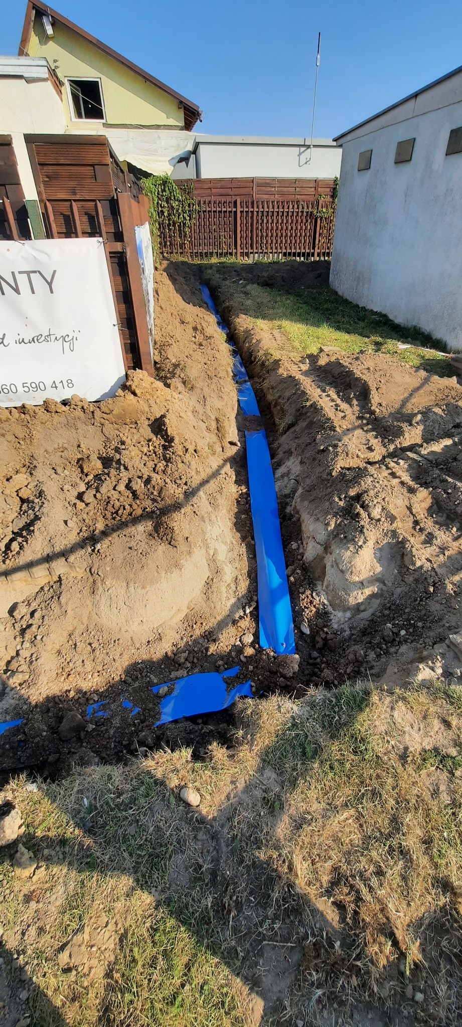 Wykopany rów z niebieską folią zabezpieczającą, prawdopodobnie przygotowany do ułożenia instalacji podziemnej, widoczne fragmenty budynku w tle.
