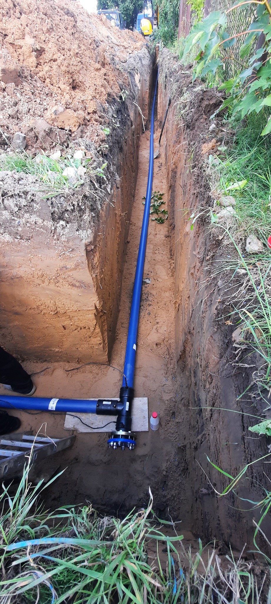 Głęboki wykop w ziemi z ułożonym niebieskim rurociągiem z czarnymi złączkami i czujnikami, widoczne ślady pracy ziemnych, w tle trawa i pojazdy.