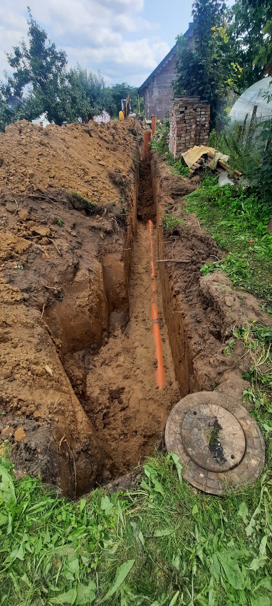 Wykonany wykop pod rury kanalizacyjne z widocznym pomarańczowym rurociągiem i studzienką kanalizacyjną na trawie, w tle budynek i koparka.
