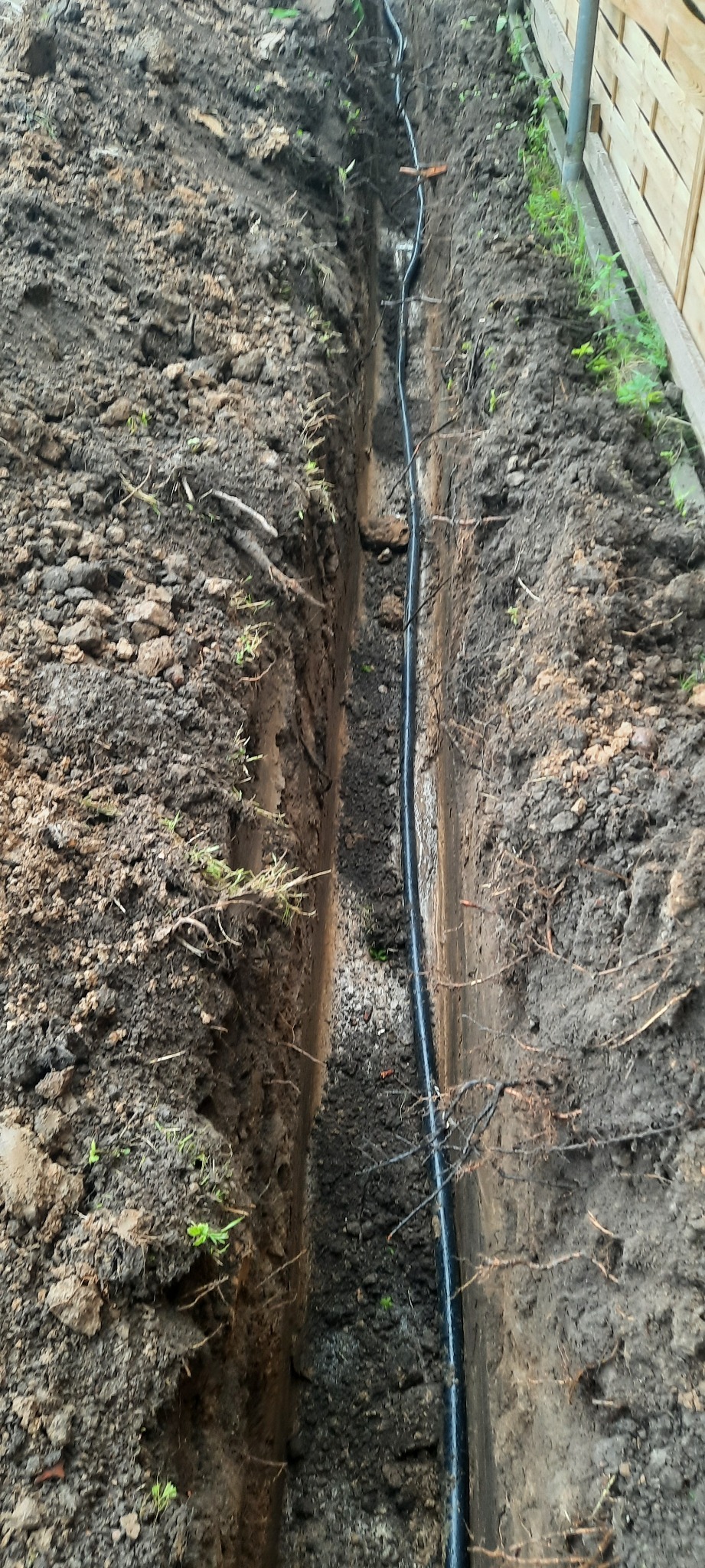 Wykopany wąski rów w ziemi z ułożonym czarnym kablem wewnątrz, widoczny fragment drewnianego ogrodzenia w tle.