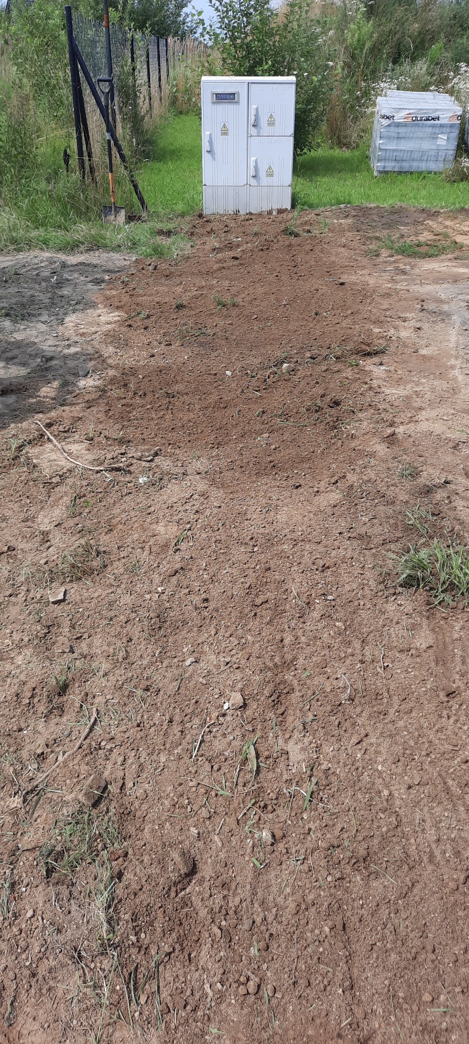 Przygotowany teren pod kostkę brukową z widoczną studzienką telekomunikacyjną i paletą kostki w tle.