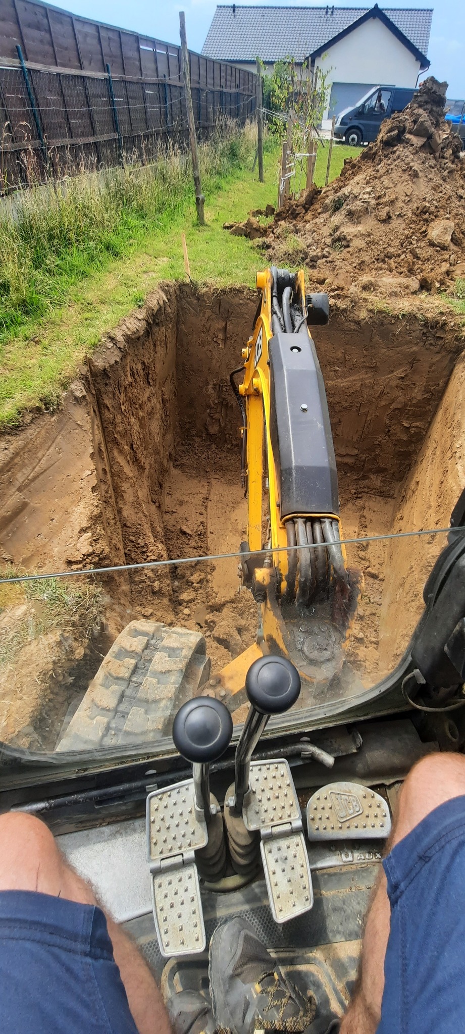 Widok z kabiny żółtej koparki JCB podczas wykopywania głębokiego, kwadratowego dołu w ziemi na trawiastym terenie z widocznym ogrodzeniem i domem w tle.