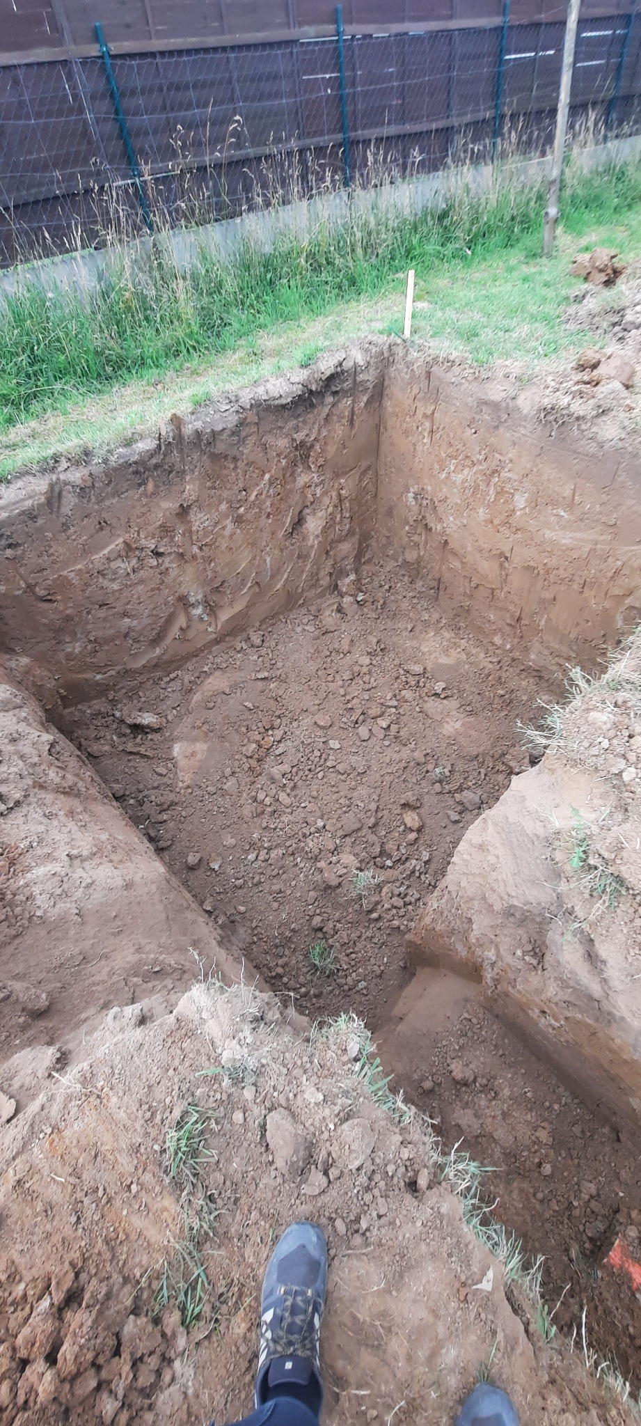 Wykopany dół w ziemi z widocznymi ścianami z gliny i ziemi, częściowo zasypany gruzem, obok rosną trawy, widoczne buty pracownika.