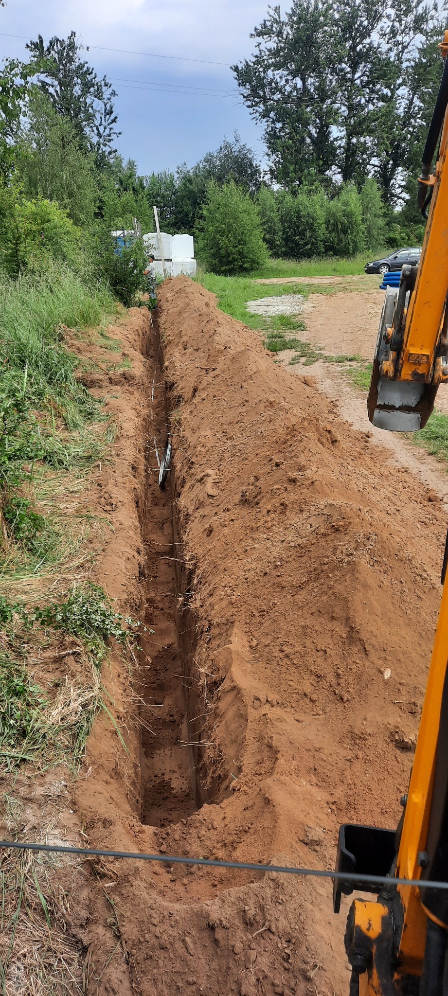 Wykopany rów w ziemi z ułożonymi rurami kanalizacyjnymi, widoczna koparka i teren budowy w tle.