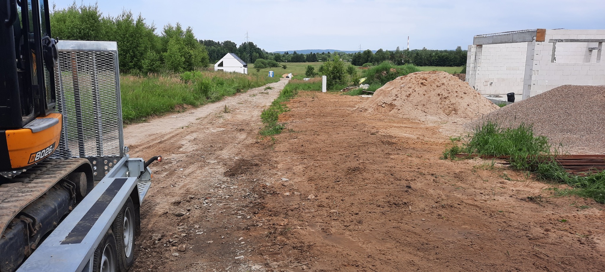 Widok placu budowy z ziemią, piaskiem i żwirem, w tle częściowo wybudowany budynek z białych bloczków oraz maszyna budowlana z przyczepą.