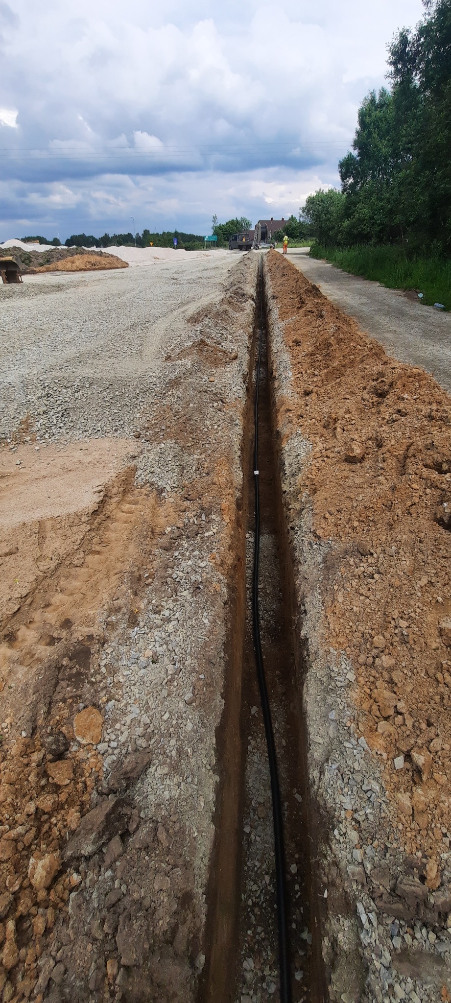 Wykop w ziemi z ułożonym kablem, widok z góry, w tle plac budowy i pochmurne niebo.