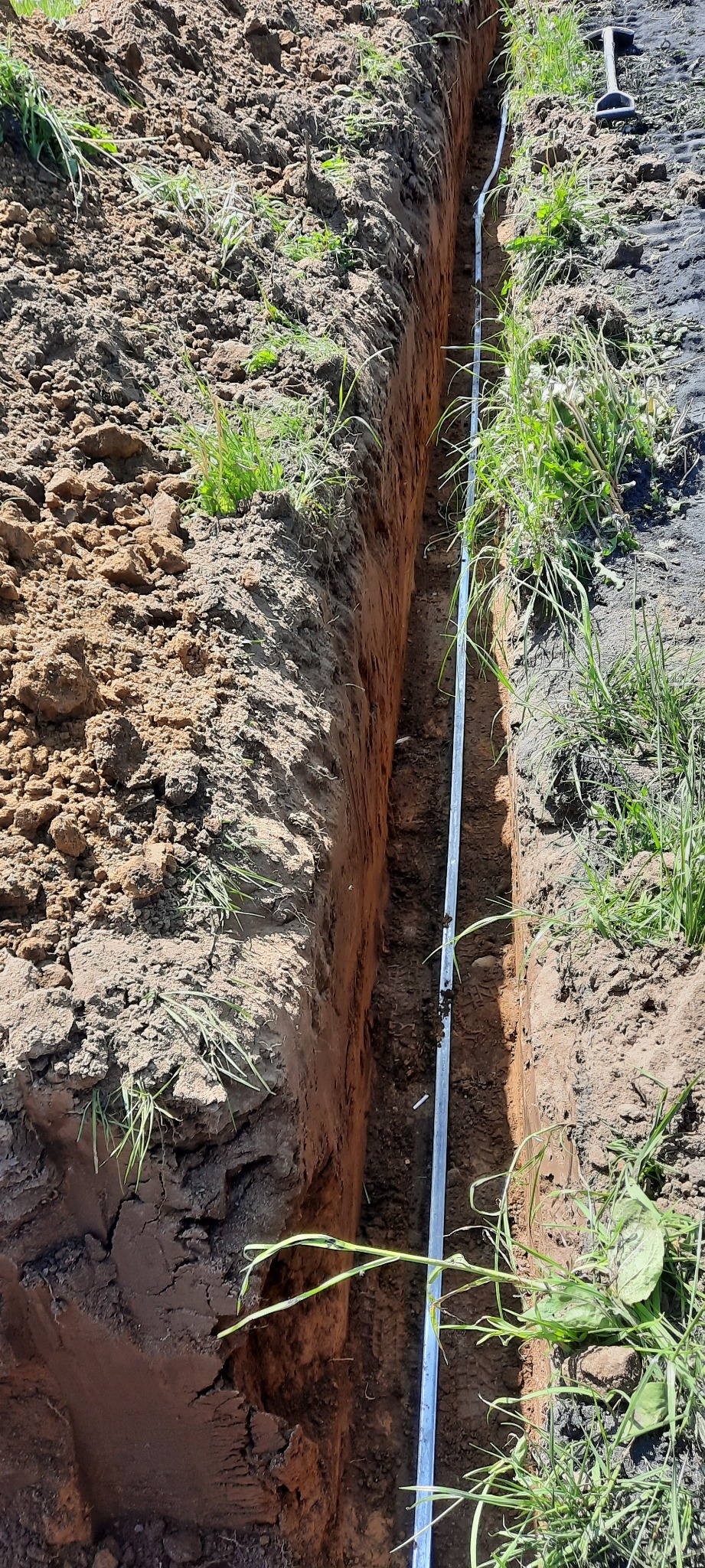 Długi, wąski wykop w ziemi z ułożoną metalową rurą na dnie, widoczne ślady opon na powierzchni ziemi po prawej stronie.