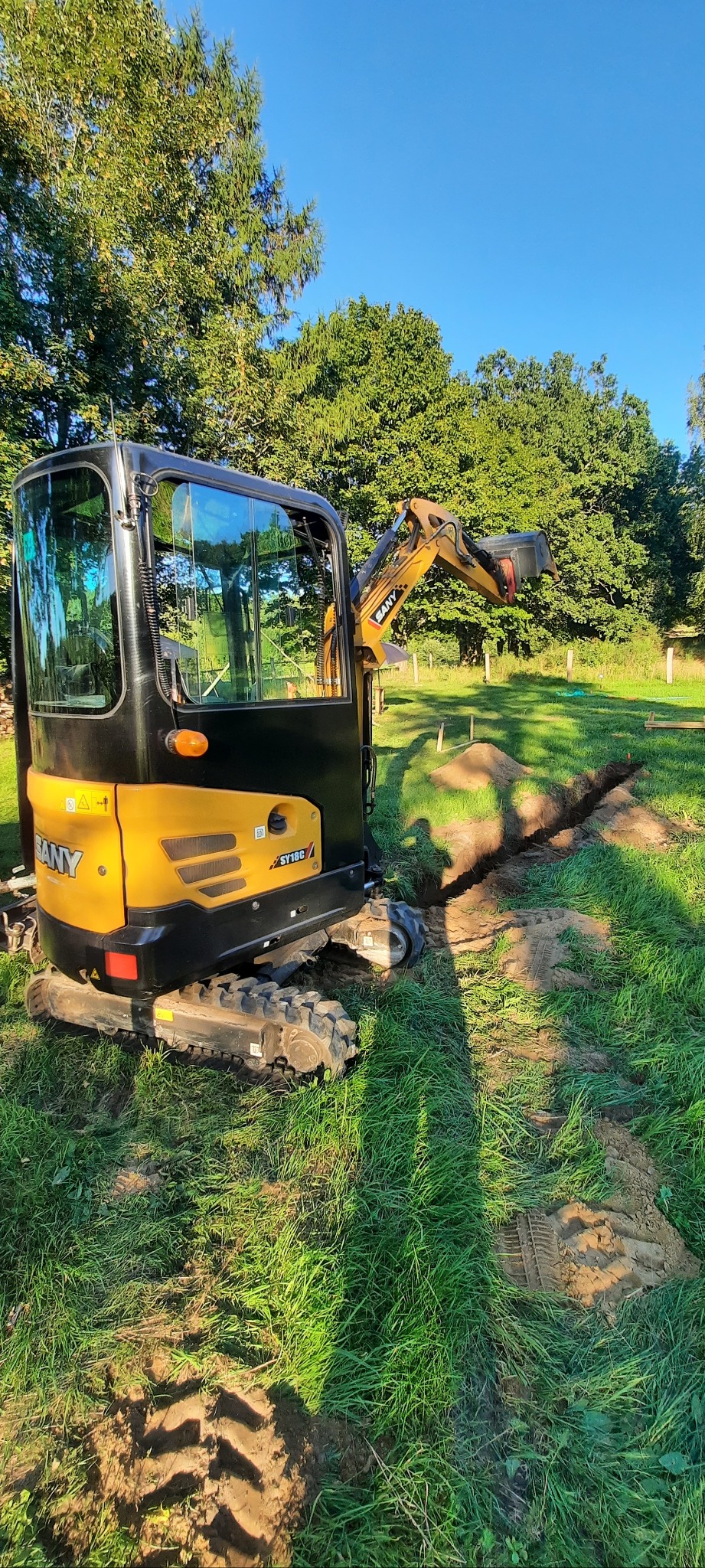 Żółto-czarna mini koparka SANY SY18C w trakcie pracy nad wykopem w zielonej trawie, z drzewami w tle i błękitnym niebem.