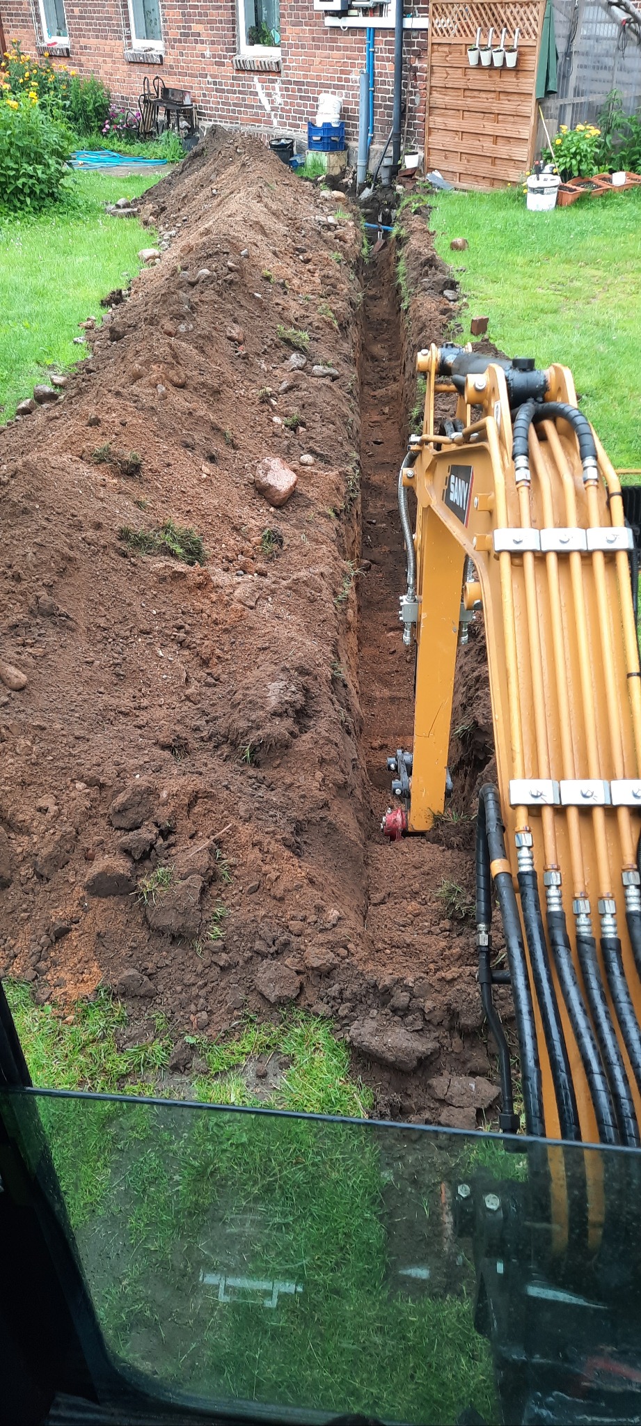 Żółta koparka SANY w trakcie wykonywania wykopu pod instalacje na działce z trawnikiem i budynkiem w tle. Widok z kabiny operatora.