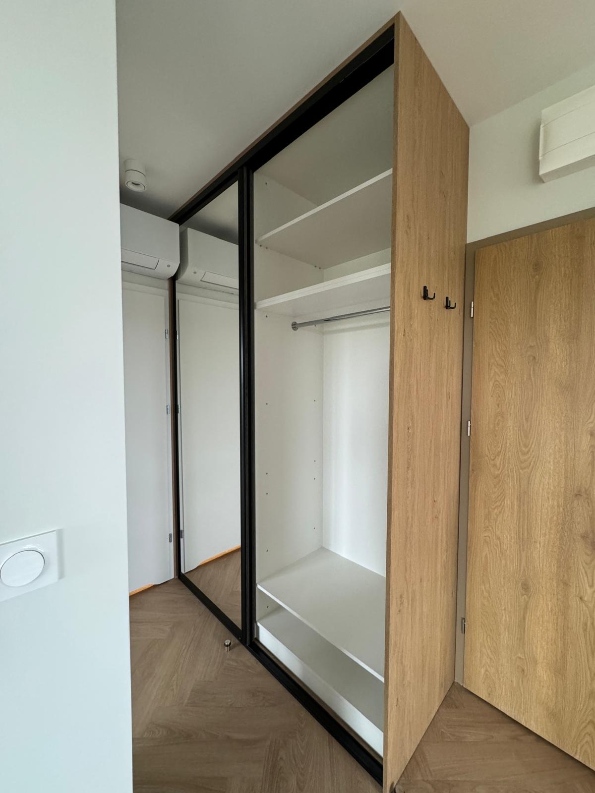 Wnętrze szafy wnękowej z przesuwnymi drzwiami z czarnymi ramami, wykończonej okleiną drewnopodobną w odcieniach dębu, z widocznymi półkami i drążkiem na ubrania, zamontowanej w pomieszczeniu...