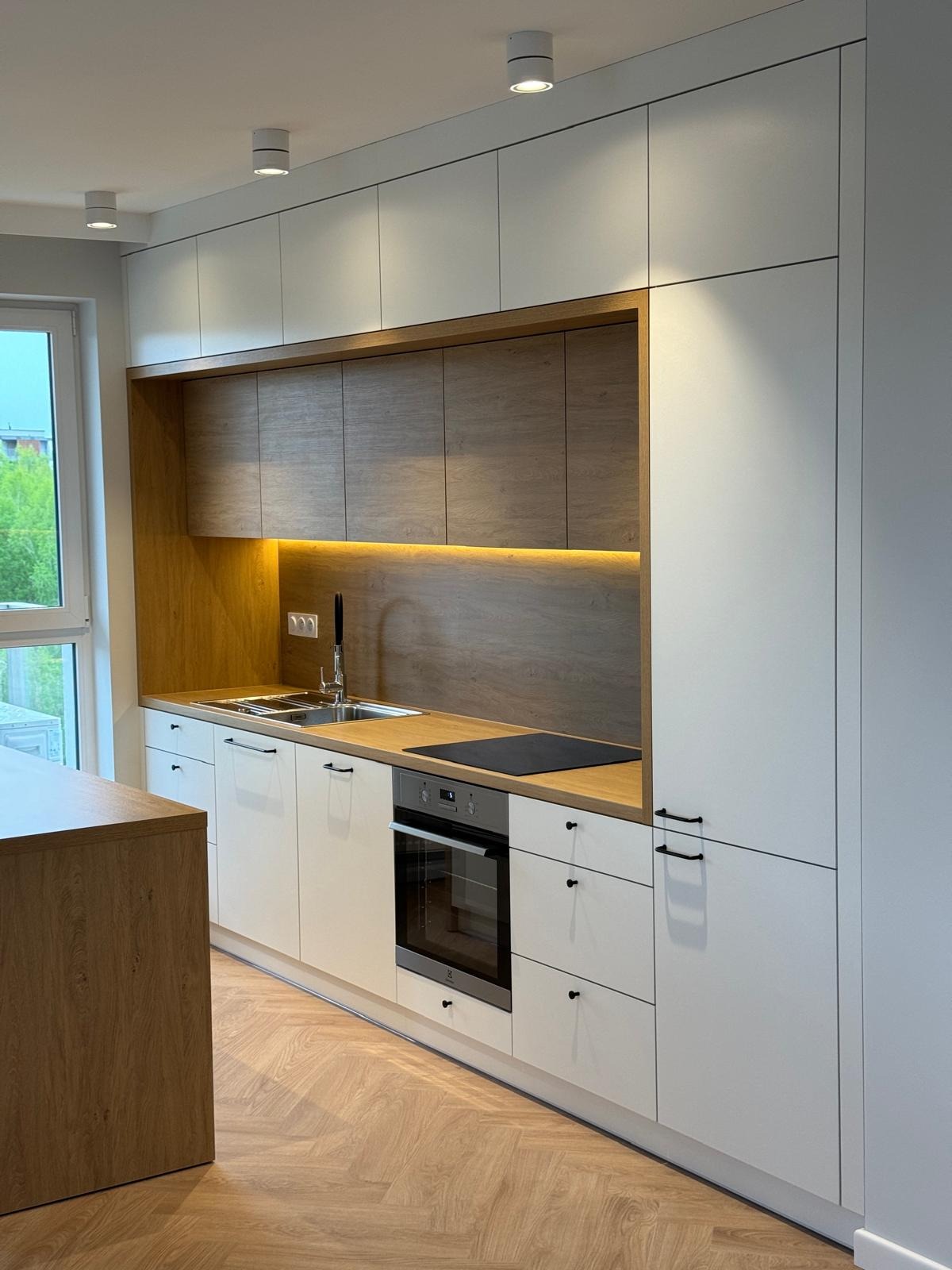 Nowoczesna kuchnia na wymiar z białymi frontami i drewnianymi akcentami, podświetleniem LED nad blatem i parkietem ułożonym w jodełkę.