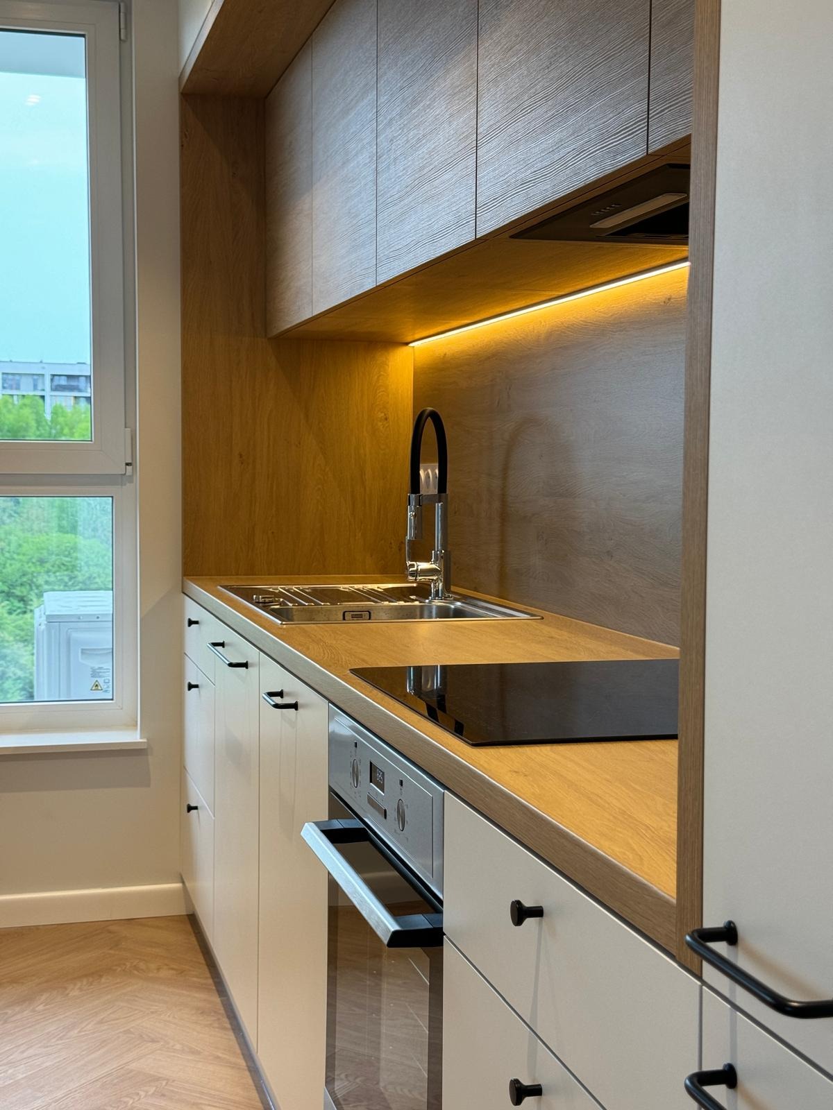 Nowoczesna kuchnia z drewnianym blatem, białymi szafkami z czarnymi uchwytami, oświetleniem LED podszafkowym i widokiem na zieleń za oknem.