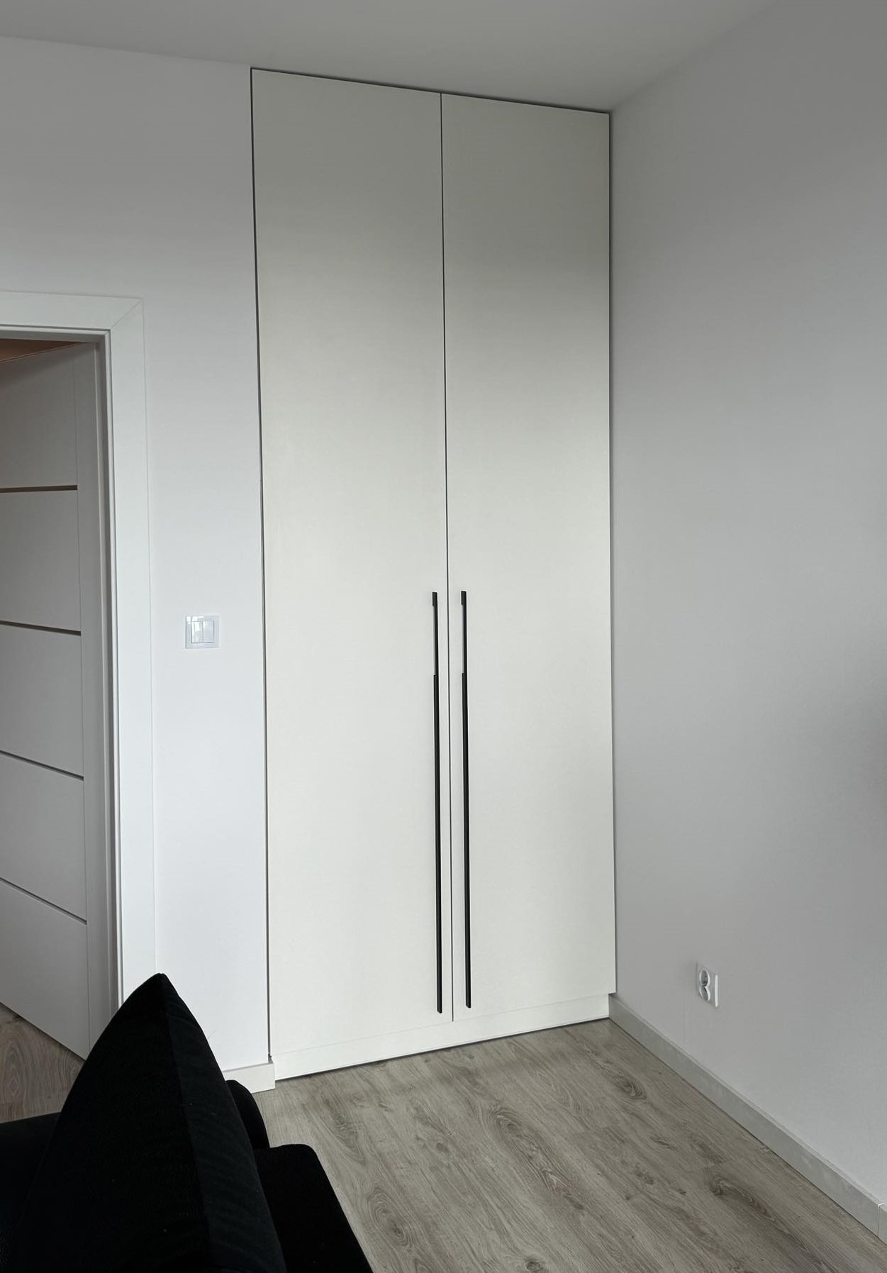Wysoka, biała szafa wnękowa z czarnymi, pionowymi uchwytami, widoczna w rogu pomieszczenia z jasnymi ścianami i panelami podłogowymi, obok drzwi z widocznymi poziomymi liniami.