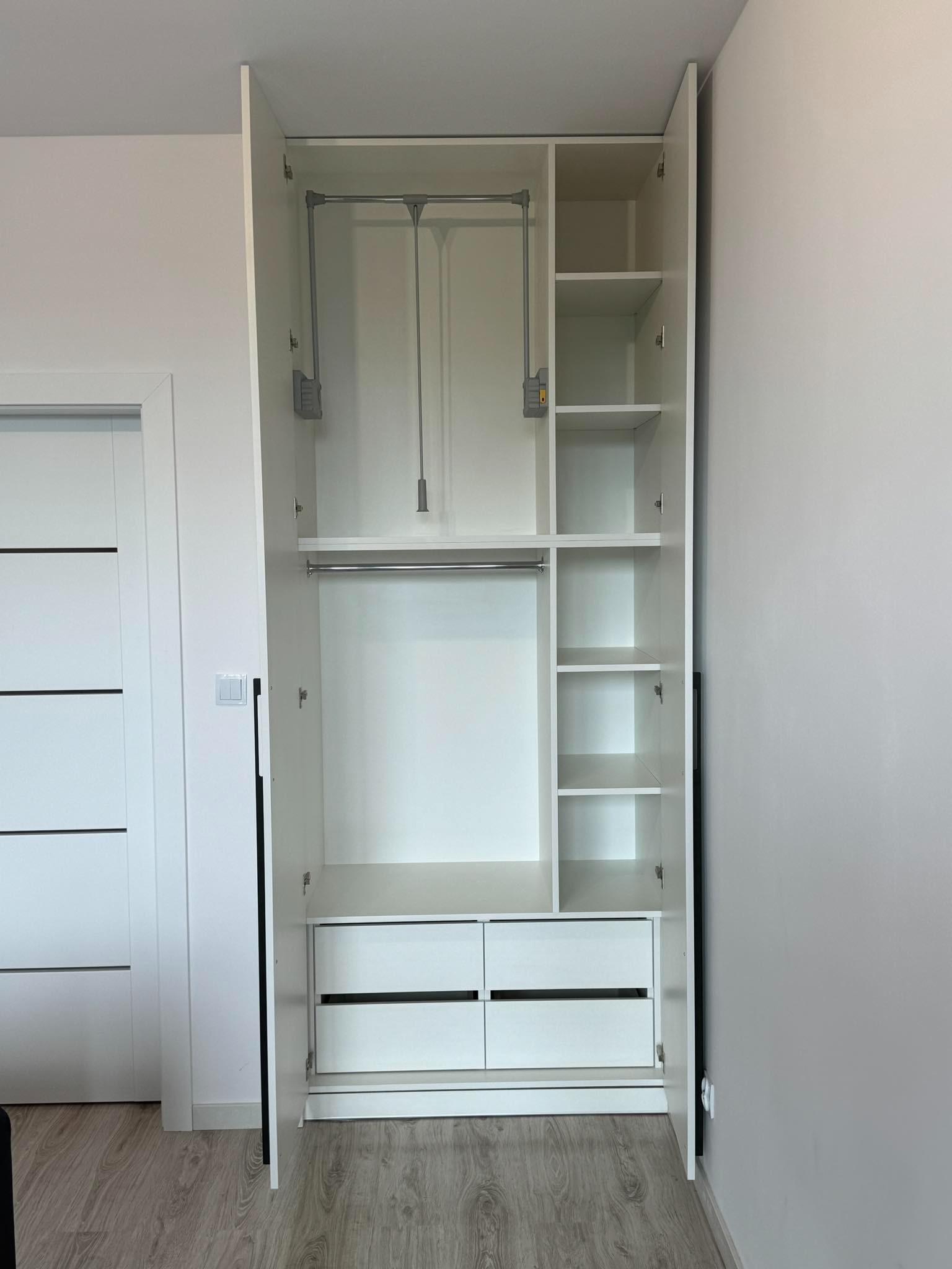 Wnętrze białej szafy wnękowej z drążkami na ubrania, półkami i szufladami, widok z otwartymi drzwiami, minimalistyczny design.