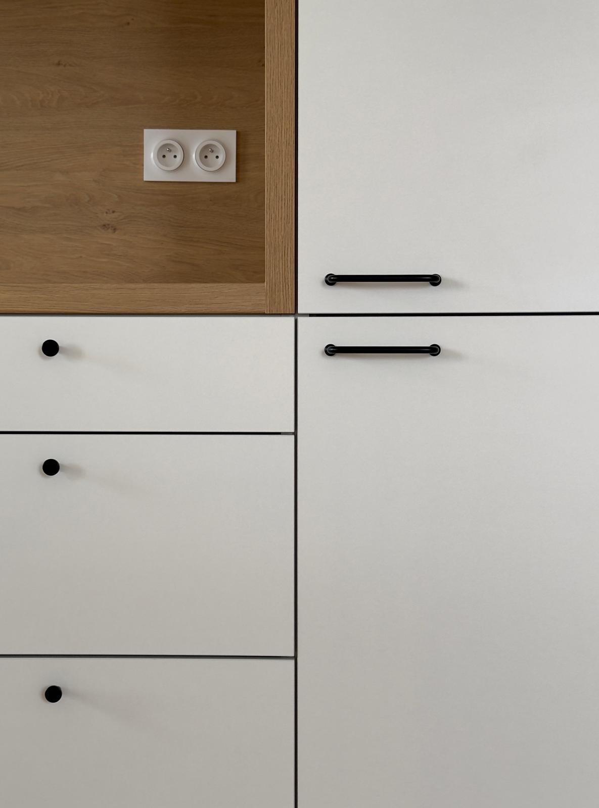 Fragment zabudowy kuchennej z białymi frontami, czarnymi uchwytami i drewnianym panelem z gniazdkiem elektrycznym.