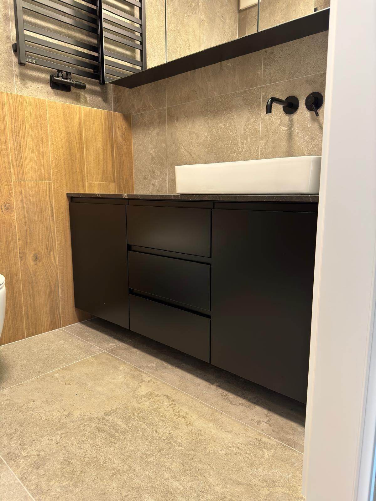 Nowoczesna łazienka z czarną szafką pod umywalkę, nablatowa umywalka, czarne baterie ścienne i grzejnik drabinkowy na tle płytek imitujących drewno i kamień.