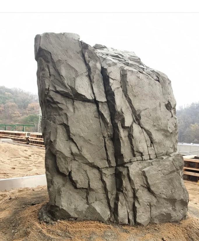 Duży, sztuczny kamień z wyraźnymi pęknięciami i nierówną fakturą, umieszczony na placu budowy z widocznymi elementami konstrukcyjnymi w tle.