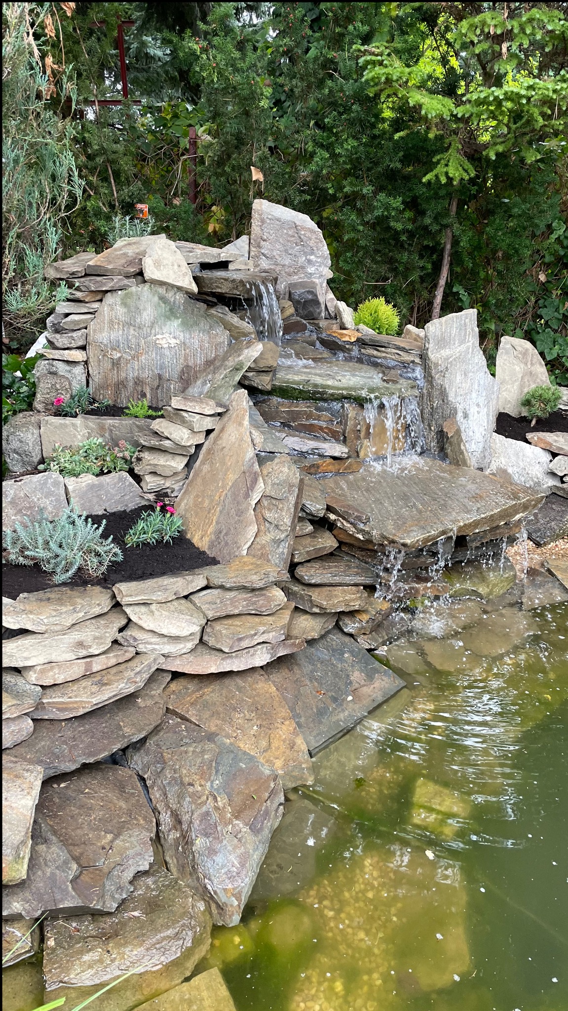 Kaskada wodna z naturalnego kamienia w ogrodzie, z roślinnością i zielonym stawem u podnóża, woda spływająca po płaskich kamieniach.