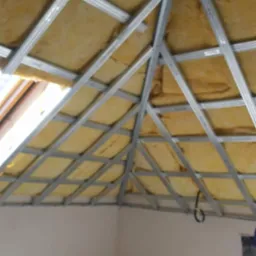 Skośny sufit poddasza z widoczną izolacją z wełny mineralnej i metalową konstrukcją szkieletową przygotowaną pod montaż płyt gipsowo-kartonowych, widok od dołu.