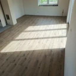 Ukończona podłoga z płytek imitujących drewno w jasnym, nowym pomieszczeniu z oknem. Płytki ułożone w regularny wzór, widoczne promienie słoneczne.