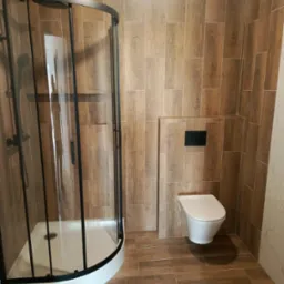 Wykończona łazienka z kabiną prysznicową typu walk-in z czarnymi ramami i toaletą podwieszaną. Płytki imitujące drewno na ścianach i podłodze.
