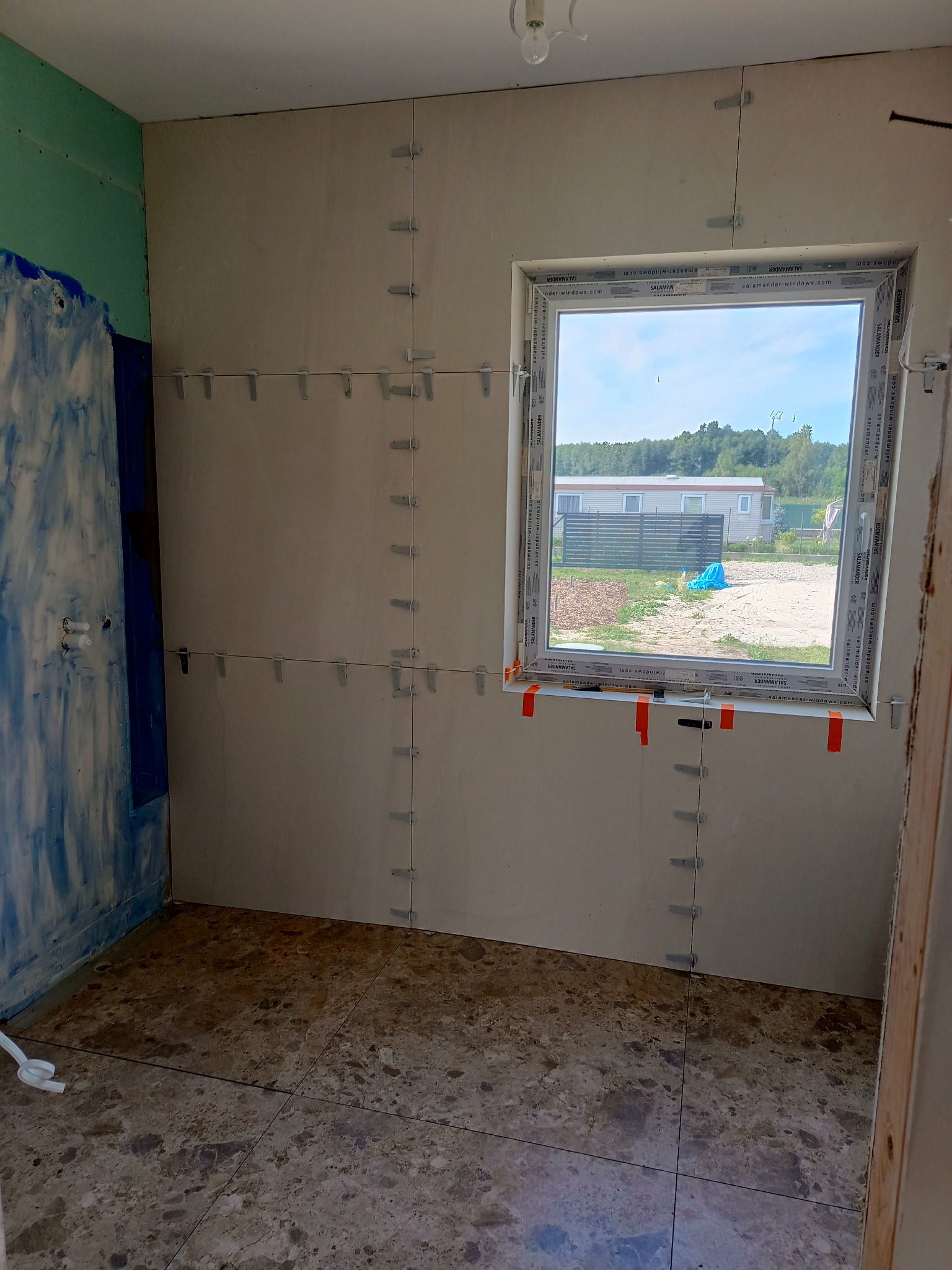 Łazienka w trakcie układania płytek ściennych, widoczne klipsy poziomujące, fragment ściany z przygotowaną hydroizolacją, marmurowe płytki podłogowe i okno z widokiem na zewnątrz.