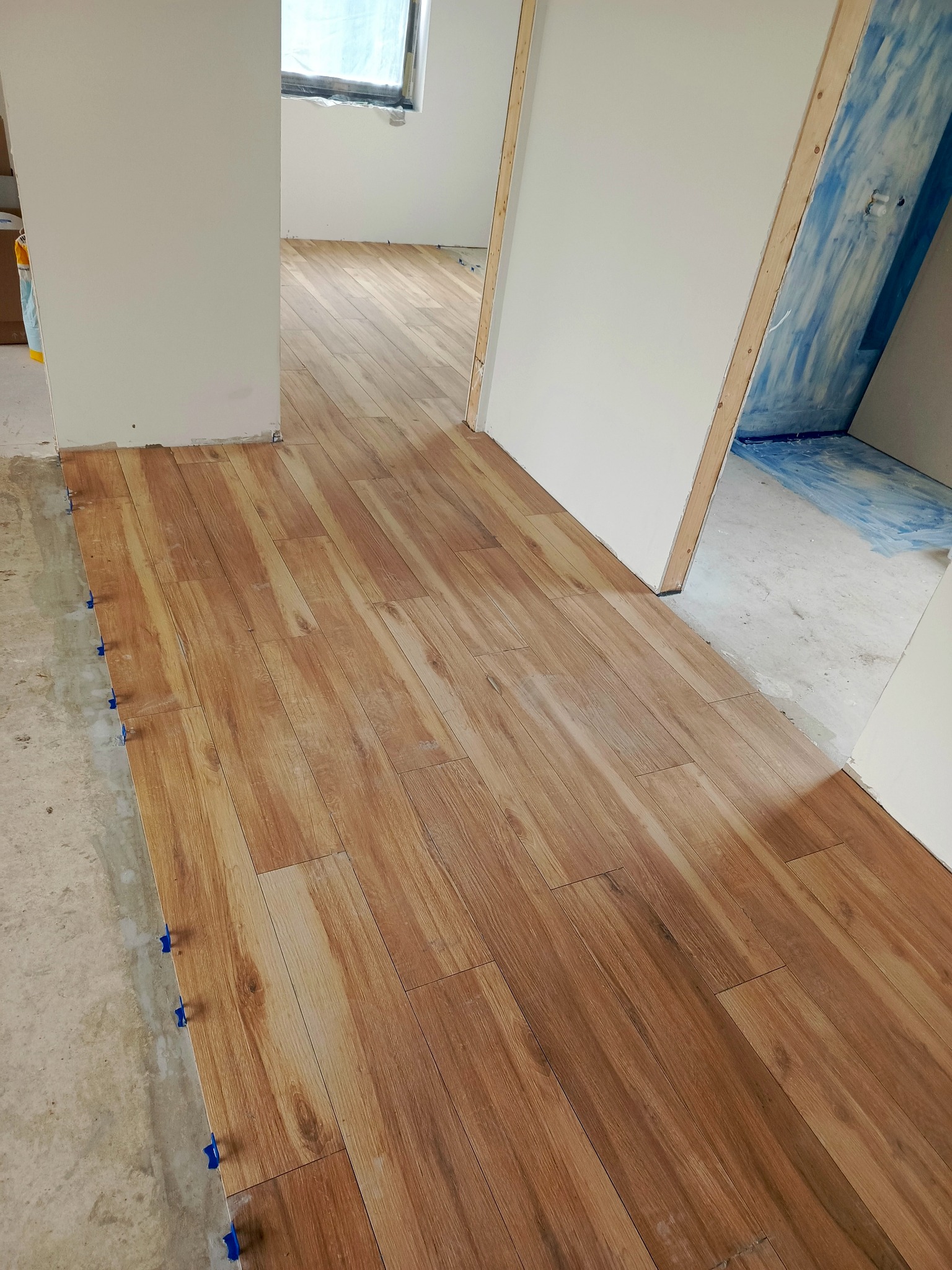 Układanie drewnopodobnych płytek podłogowych w nowym mieszkaniu, z widocznymi niebieskimi krzyżykami dystansowymi i surowym podłożem w tle.